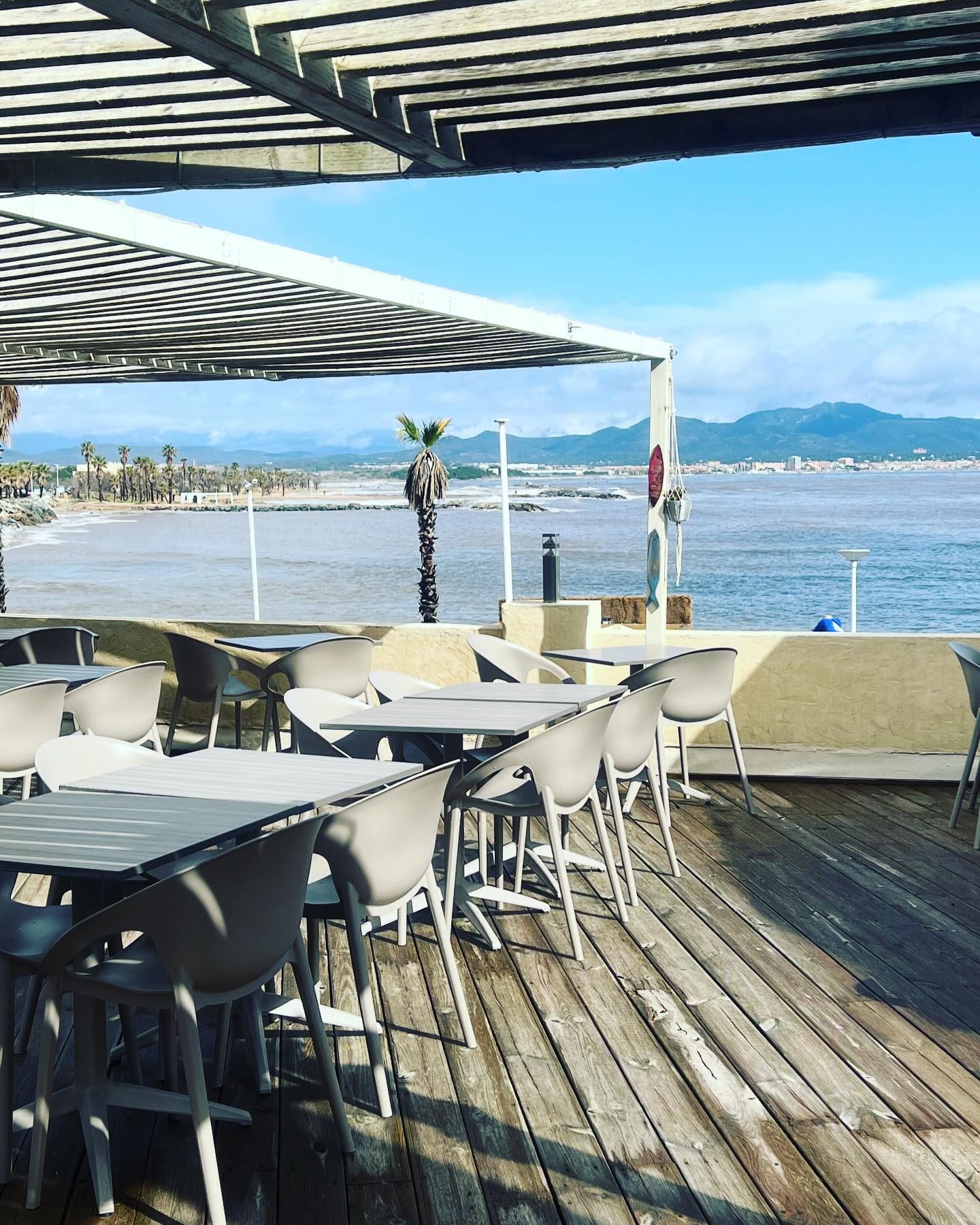 La terrasse sous pergola, face à la plage et à la baie, avec ses tables grises et ses fauteuils clairs